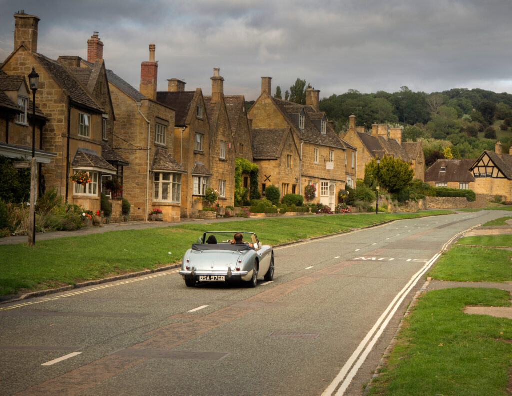 silver old fashioned classic car driving through the Cotswolds. Views of the Cotswold golden stone.