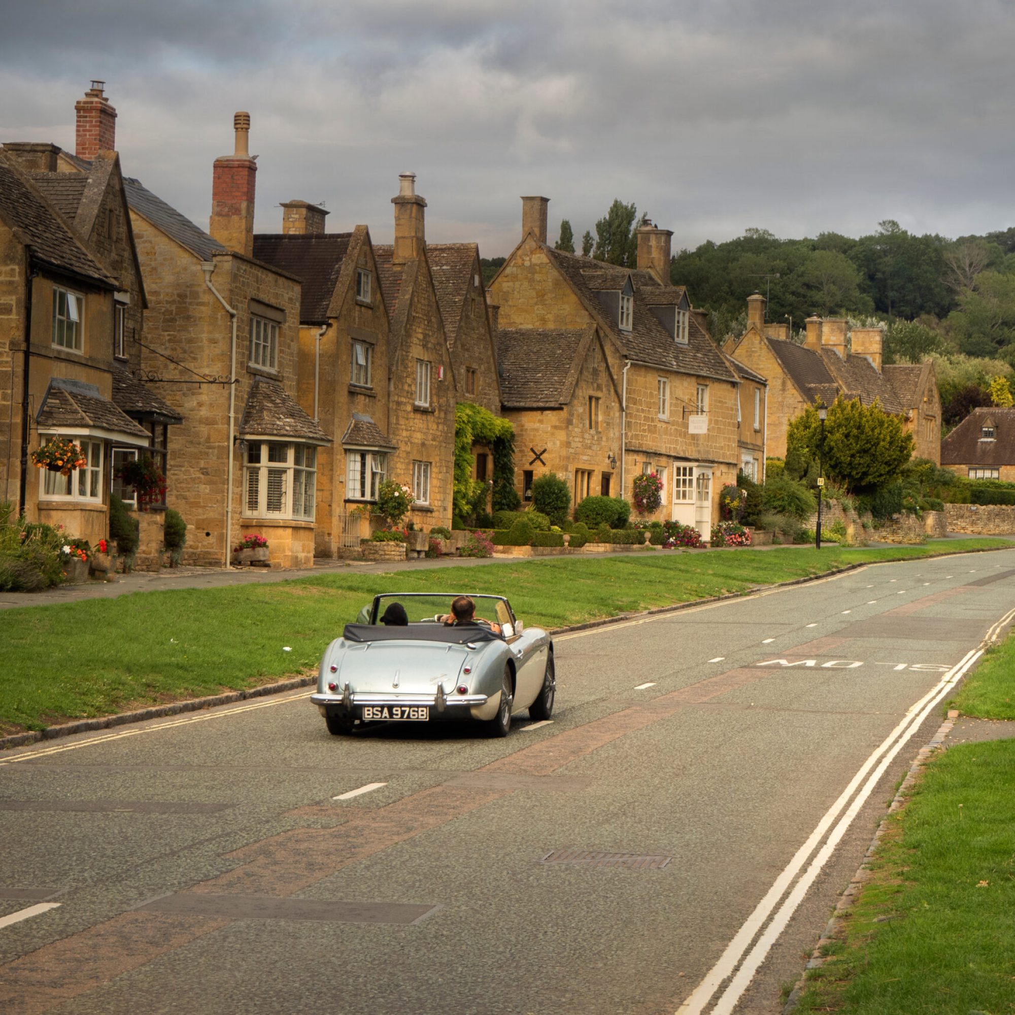 silver old fashioned classic car driving through the Cotswolds. Views of the Cotswold golden stone.