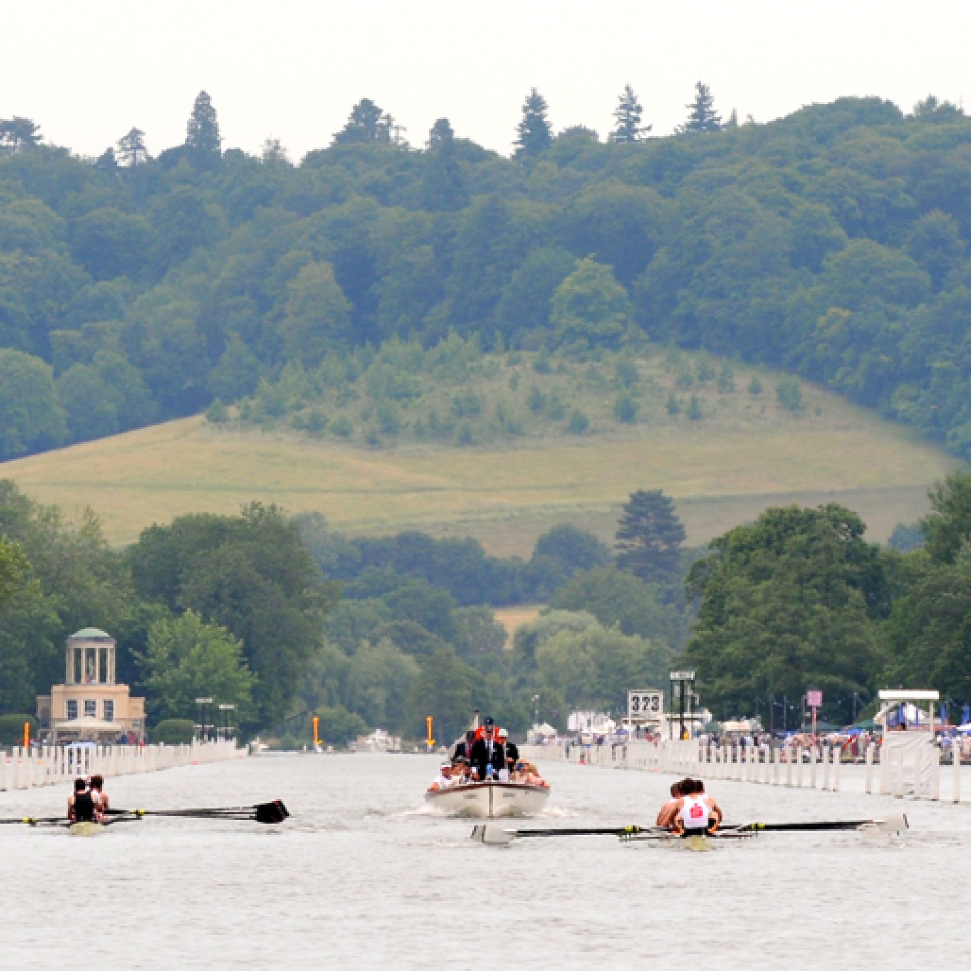 In the foreground there are two rowing boats rowing down the River Thames at Henley Royal Regatta. In the background is Temple Island and green rolling hills.